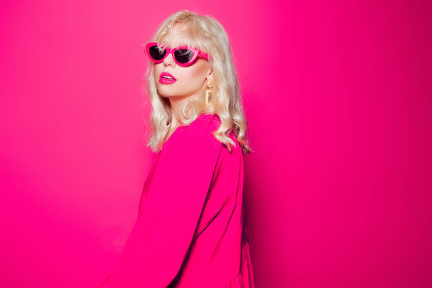 Blondýna pózuje na růžovém pozadí ve stylových růžových brýlích a šatech. Dívka model pózování, světlé fotografie móda a styl, zlaté šperky 80. a 90. léta. - Fotografie, Obrázek