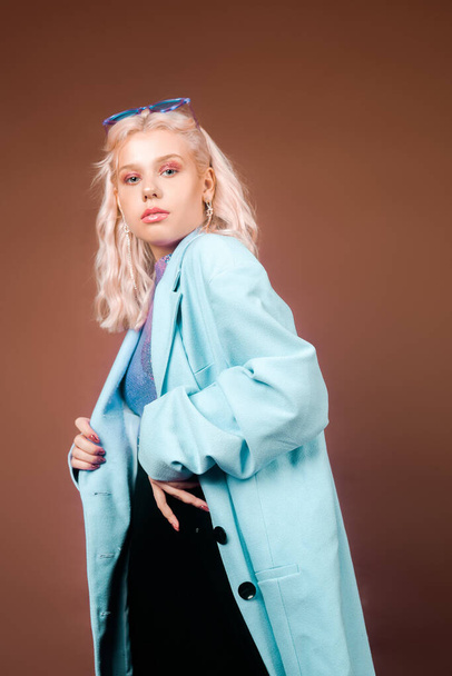 Blondi turkoosi takki, sininen takki paljetteja, poseeraa studiossa ruskealla taustalla. Huippumuotia, 90-80-lukua. Tyttö tanssii ja liikkuu. Malli vaaleanpunainen meikki. - Valokuva, kuva