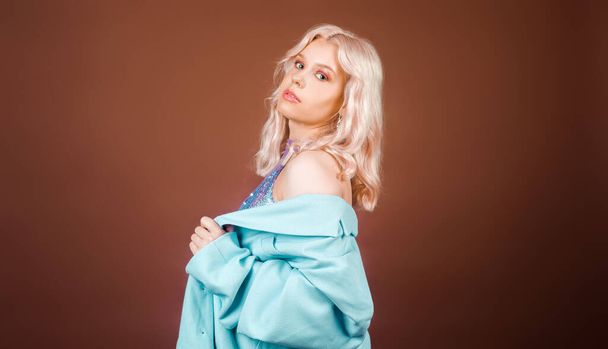 Eine Blondine in türkisfarbener Jacke, in blauer Jacke mit Pailletten posiert im Studio vor braunem Hintergrund. High Fashion, 90er, 80er. Das Mädchen tanzt und bewegt sich. Model mit rosa Make-up. - Foto, Bild