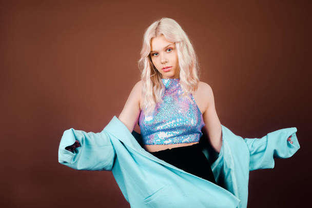 A blonde in a turquoise jacket, in a blue jacket in sequins, poses in the studio on a brown background. High fashion, 90s 80s. The girl dances and moves. Model with pink makeup. - Foto, immagini