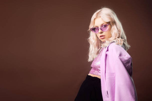 Блондинка в рожевій куртці в окулярах, піджак з блискітками позує в студії на коричневому фоні. Висока мода, 90-ті 80-ті роки. Дівчина танцює і рухається. Модель
 - Фото, зображення