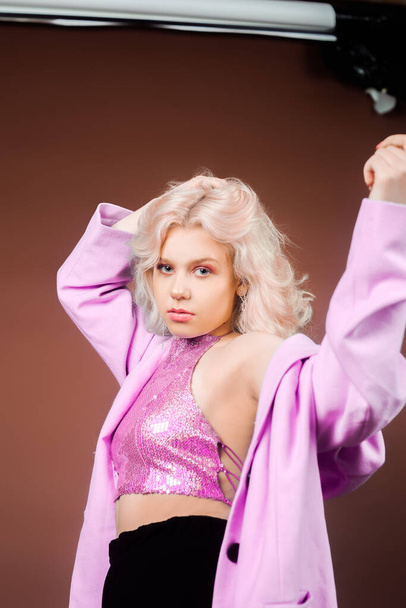 A blonde woman in a pink jacket, a jacket in sequins poses in the studio on a brown background. High fashion, 90s 80s. The girl dances and moves. Model with pink makeup - Fotoğraf, Görsel
