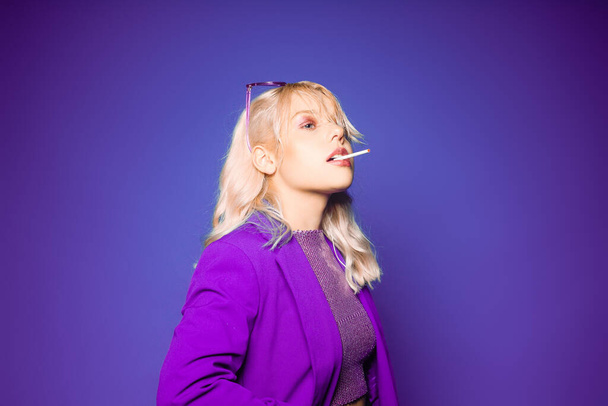 Incredibly fashionable blonde woman in a purple jacket and glasses in the style of 80s 90s poses. On a purple background. Fashionable woman smoking - Photo, Image