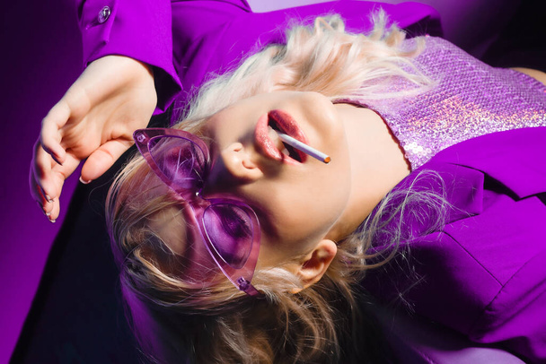Incredibly fashionable blonde woman in a purple jacket and glasses in the style of 80s 90s sits on an armchair and poses. On a purple background. Fashionable woman smoking. - Photo, Image