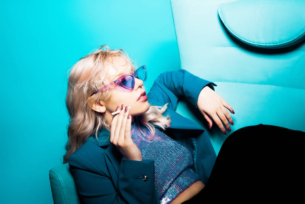 Niesamowicie modna blondynka w niebieskiej kurtce i okularach w stylu lat 80., siedzi na fotelu i pozuje. Na niebieskim tle. Modna kobieta paląca. - Zdjęcie, obraz