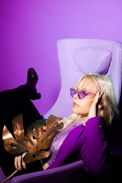 Девушка в фиолетовой куртке и фиолетовых очках сидит на стуле. Модное фото в стиле 80-х. - Фото, изображение