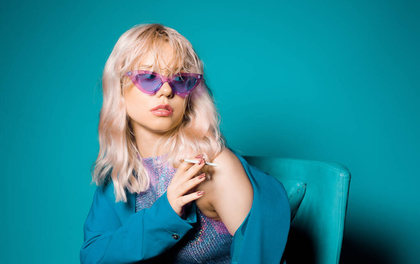 Incredibly fashionable blonde woman in a blue jacket and glasses in the style of 80s 90s sits on an armchair and poses. On a blue background. Fashionable woman smoking. - Fotografie, Obrázek