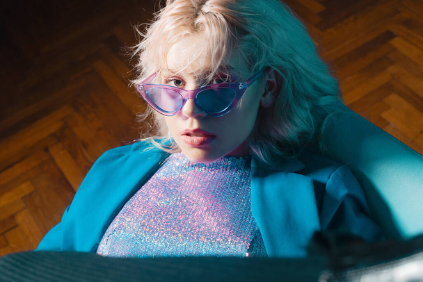 A lány benne van a 80-as évek 90-es évekbeli klipjében. Rövid szőke frizura. A lány egy széken ül kék kabátban, fényes kabátban, szemüvegben. Divat & stílus - Fotó, kép