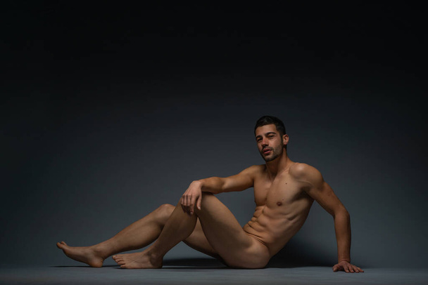 Herrliches nacktes männliches Modell sitzt isoliert auf dem Boden in einem Studio und schaut in die Kamera - Foto, Bild