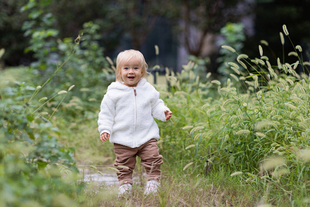 Portrait of cute little caucasian baby girl one year old takes the first steps and learns to walk in park at autumn. Stylish infant with blonde hair and blue eyes wearing fashionable clothing. Happy - Fotó, kép
