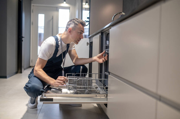 Man squatting peeking into dishwasher in kitchen - Foto, afbeelding