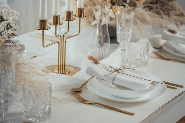 Dinner Party at Home: Nahaufnahme eines dekorierten Tisches mit Tellern, Kristallgläsern, Servietten, goldenem Essgeschirr, Kerzenständern mit Kerzen und getrocknetem Blumengesteck in einer Vase. - Foto, Bild