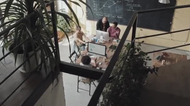 Hoge hoek van vier multi-etnische collega 's die gebruik maken van computers, overdag aan tafel zitten in een loft-achtige cowerkruimte, aan het project werken - Video