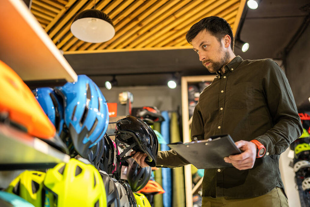 Bike shop manager controleert helm prijsinformatie op tablet, verkoper maakt een inventaris in de sportwinkel. Thema van kleine bedrijven die fietsen verkopen. Verkoper in handen document checklist in fietsenwinkel. - Foto, afbeelding