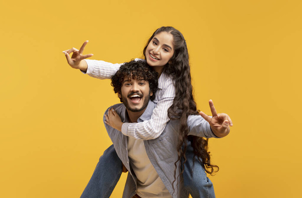 pareja india romántica divirtiéndose juntos, mostrando gesto V-signo, chico a cuestas su novia, fondo amarillo - Foto, imagen