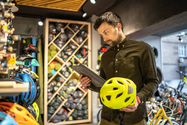 Un responsable de magasin de vélo fait un inventaire des casques de sport dans un magasin de vélo. Le propriétaire d'un magasin de sport avec un presse-papiers dans ses mains vérifie les prix des casques de vélo dans la vitrine. - Photo, image