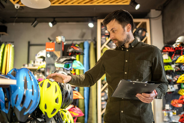 Um gerente de loja de bicicletas masculino faz um inventário de capacetes esportivos em uma loja de bicicletas. O proprietário de uma loja de esportes com uma área de transferência em suas mãos verifica os preços dos capacetes de bicicleta na vitrine. - Foto, Imagem