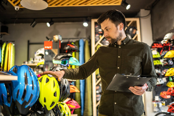 Bike shop manager checks helmet price information on tablet, seller makes an inventory in sports shop. Theme of small business selling bicycles. Seller in hands document checklist in bicycle store. - Photo, Image