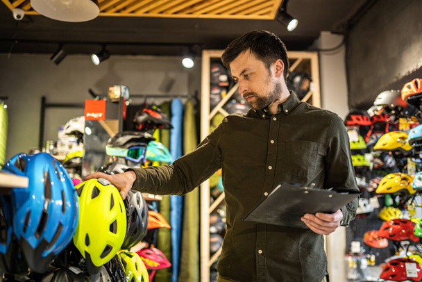 Um gerente de loja de bicicletas masculino faz um inventário de capacetes esportivos em uma loja de bicicletas. O proprietário de uma loja de esportes com uma área de transferência em suas mãos verifica os preços dos capacetes de bicicleta na vitrine. - Foto, Imagem