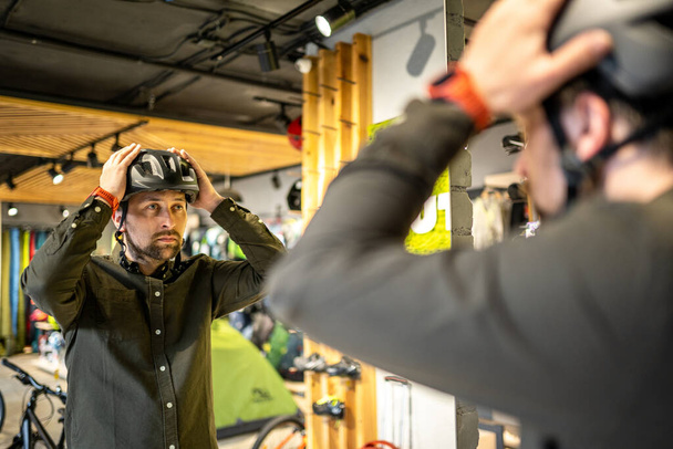 Běloch si zkouší černou cyklistickou helmu u zrcadla v obchodě se sportovním zbožím. Muž kupující si vybere bezpečnostní helmu pro cyklistiku. Nakupování v obchodě s koly. Osoba si upraví řemínek sportovní přilby. - Fotografie, Obrázek