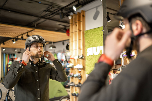 Мужчина осматривает велосипедные шлемы в спортивном магазине, глядя на себя в зеркало. Пробую новый спортивный шлем в магазине велосипедов. Мужчина надевает велосипедный шлем в магазине. Клиент в магазине велосипедов примеряет велосипедный шлем. - Фото, изображение