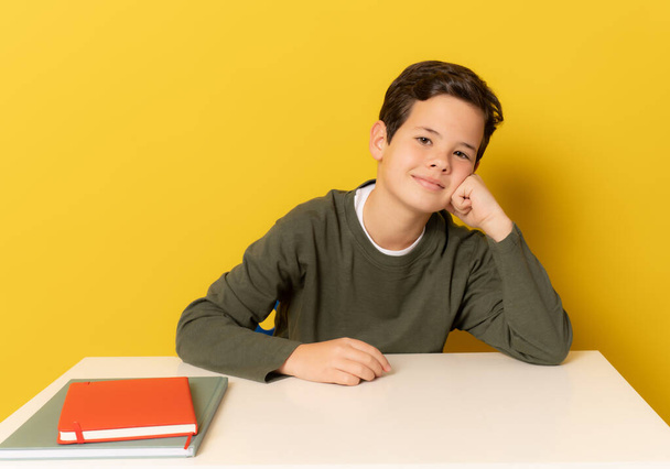 Sorridente studente ragazzo seduto sulla scrivania isolato su sfondo giallo. - Foto, immagini