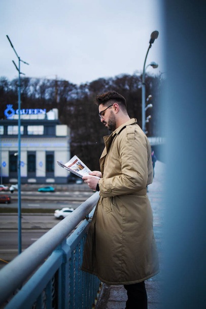 Young bearded man reads the latest news. Confident young man in winter coat reading newspaper while standing outdoors on bridge with cityscape in background. - Foto, Imagem