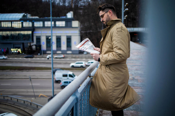 若い髭の男が最新のニュースを読んでいる。裏の街並みと橋の上に屋外に立っている間、冬のコートを読む新聞で若い男を自信を持って. - 写真・画像