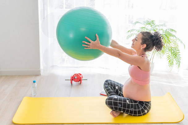 Sonriente mujer embarazada bastante joven sosteniendo una pelota de fitness verde en sus brazos sentada en una esterilla de ejercicio con una botella de agua a su lado para la hidratación. concepto de cuidado de la salud embarazo - Foto, imagen