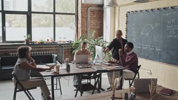 Długie ujęcie czterech wielonarodowych kolegów korzystających z komputerów, siedzących przy stole w przestrzeni współpracowników na poddaszu w ciągu dnia, pracujących nad projektem - Materiał filmowy, wideo