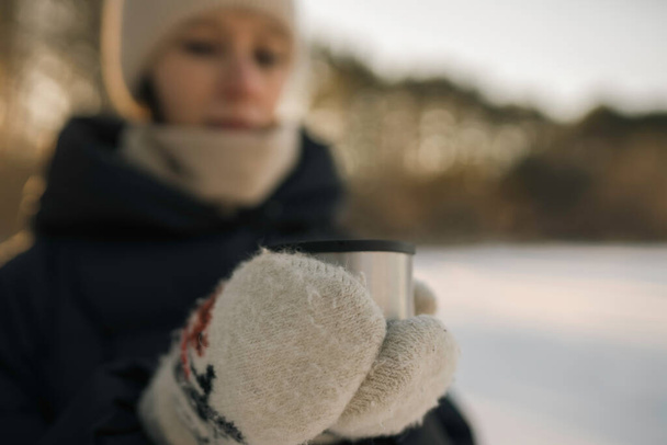 Woman with hot tea into a mug, walking in snowy frozen winter forest at sunset. Adventure, tourism and camping concept - Fotografie, Obrázek