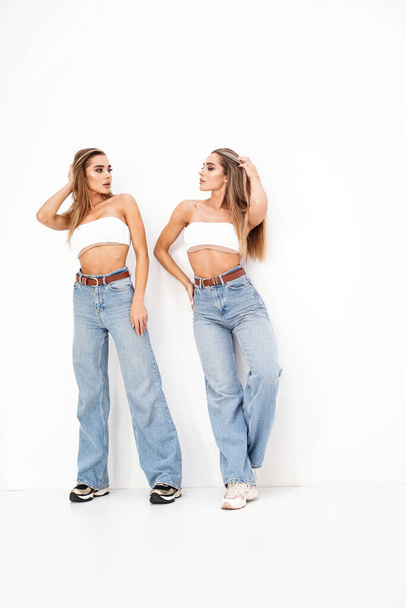 Two beautiful young women with long blond hair and glamour makeup wearing fashionable jeans, standing over white studio background. Sisters. Twins. Studio shot. Copy space - Foto, Bild
