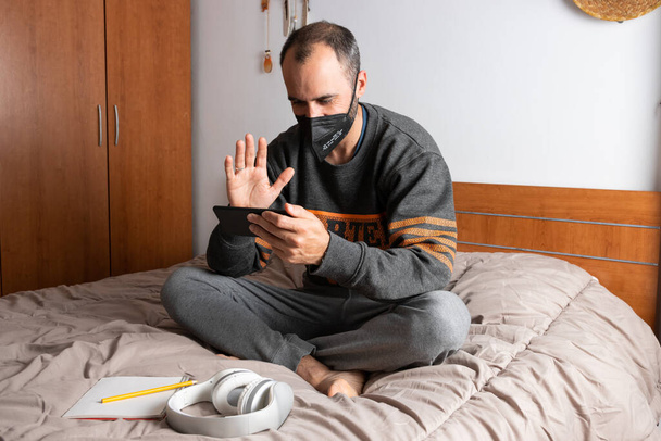 Hombre con auriculares, teléfono móvil y mascarilla, sentado en su cama y confinado a su habitación - Foto, imagen