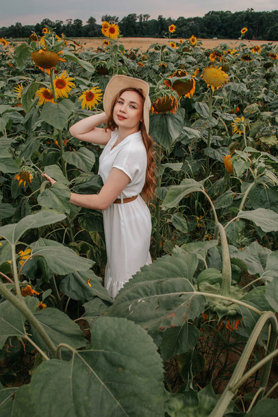 Ομορφιά χαρούμενη έφηβη κοπέλα με ηλιοτρόπιο απολαμβάνοντας τη φύση και γελώντας στο καλοκαιρινό ηλιοτρόπιο πεδίο. νεαρή κοπέλα σε ένα χωράφι με ηλιοτρόπια σε ένα λευκό φόρεμα με καπέλο. - Φωτογραφία, εικόνα