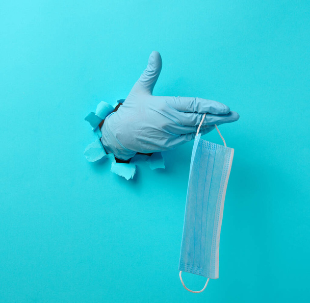 una mano femenina sostiene una máscara médica desechable para protegerse contra los virus durante una epidemia y pandemia. Parte del cuerpo sobresale de un agujero roto en papel azul - Foto, Imagen