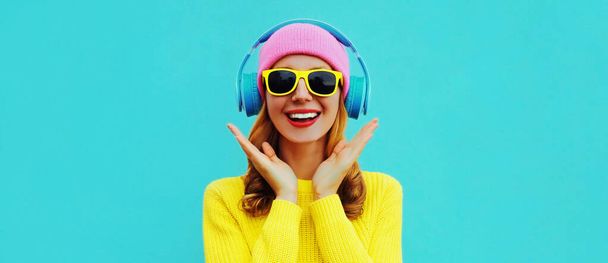 Portrait of happy surprised smiling young woman model with headphones listening to music wearing colorful yellow sweater on blue background - Zdjęcie, obraz