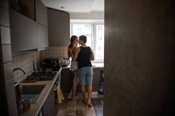 Young couple of millennials in black t-shirts, jeans hugging in the kitchen in the morning by window. Loving lovers have fun before breakfast at home. Man strokes womans hair. Lifestyle apartment life - Foto, Bild