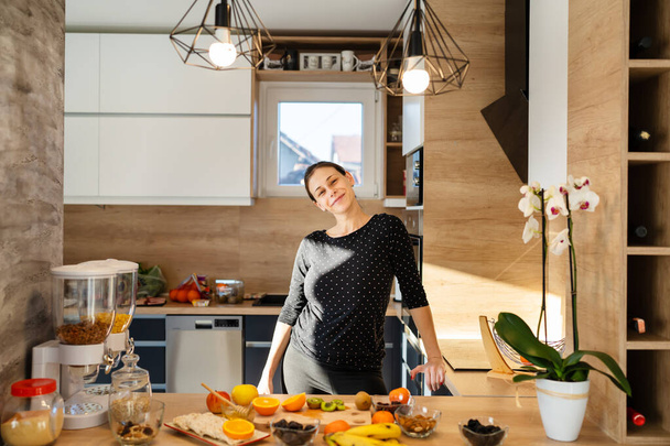 Vooraanzicht portret van een jonge vrolijke blanke vrouw in de keuken tijdens het bereiden van gezonde maaltijd ontbijt met havermout fruit sinaasappelen kiwi rozijnen en honing echte mensen duizendjarige vrouwelijke - Foto, afbeelding