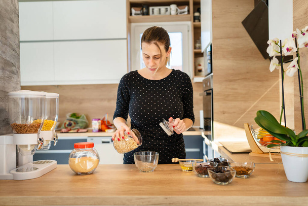 One young caucasian woman standing in the kitchen making healthy breakfast front view millennial female adult putting oatmeal oatflakes in the bowl preparing meal real people copy space - Photo, Image