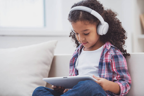 Linda niña afroamericana con ropa casual y auriculares usando una tableta y escuchando música mientras está sentada en un sofá en la habitación
. - Foto, Imagen