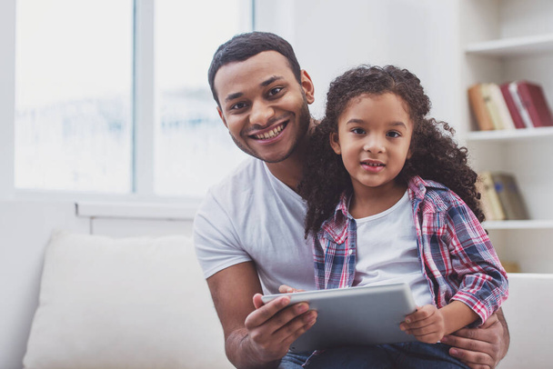 Linda niña afroamericana con ropa casual sentada en las rodillas de su padre. Ambos usando una tableta, mirando a la cámara y sonriendo
. - Foto, Imagen