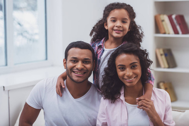 süßes kleines afroamerikanisches Mädchen und ihre schönen jungen Eltern umarmen sich, schauen in die Kamera und lächeln, während sie auf einem Sofa im Zimmer sitzen. - Foto, Bild