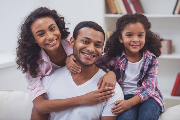 Cute little Afro-American girl and her beautiful young parents hugging, looking at camera and smiling while sitting on a sofa in the room. - Фото, зображення