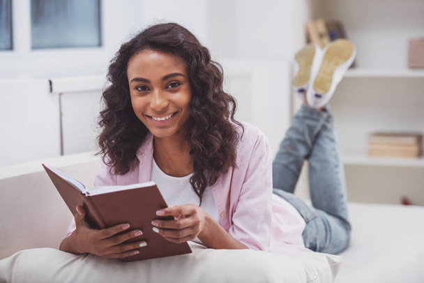Young beautiful Afro-American woman reading a book, looking at camera and smiling while lying on a sofa in the room. - Photo, Image