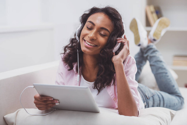 Młode piękne afro-amerykański kobieta w słuchawki za pomocą laptopa, słuchanie muzyki i uśmiechając się leżąc na kanapie w pokoju. - Zdjęcie, obraz