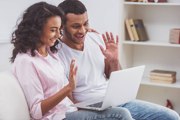 Jovem casal afro-americano bonito usando um laptop, acenando e sorrindo enquanto sentado em um sofá na sala
. - Foto, Imagem
