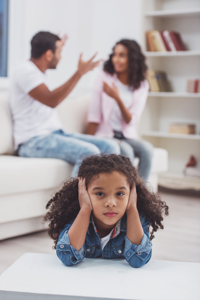 Schattig klein Afro-Amerikaanse meisje over haar oren, op de achtergrond haar ouders ruzie. - Foto, afbeelding