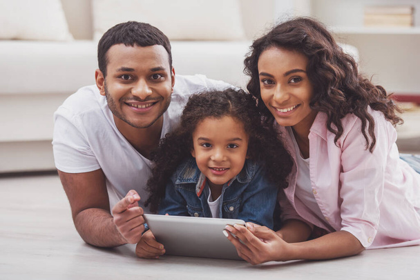 Jolie petite fille afro-américaine et ses beaux jeunes parents à l'aide d'une tablette, regardant la caméra et souriant tout en étant couché sur le sol dans la chambre
. - Photo, image