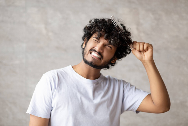 Сухие и спутанные волосы. Индийский молодой парень в белой футболке в сочетании с кистью и с болью от запутанных кудрявых волос - Фото, изображение