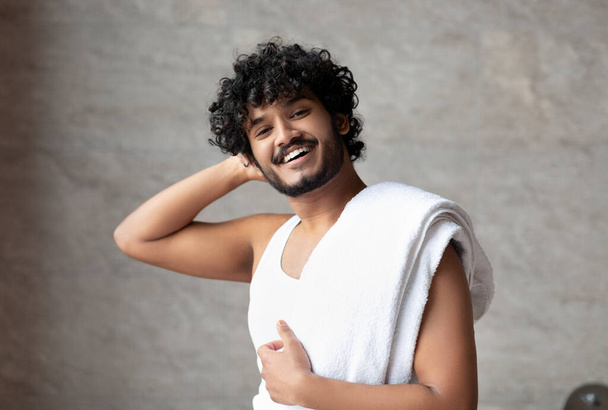 Mužská péče o krásu. Šťastný indián s ručníkem na rameni stojící v koupelně a usmívající se na kameru - Fotografie, Obrázek